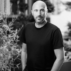 Black and white headshot of Founder Adam Rubins, stood with hands in pockets with welcoming expression.